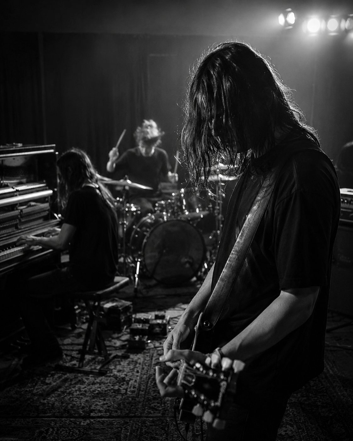 A band rehearsing in low light: guitarist in the foreground, drums and upright piano behind